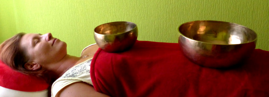 Klangmassage mit tibetischen Klangschalen – Herzschale und Beckenschale auf dem Körper während einer Klangbehandlung, tief entspannte Person auf der Liege unter roten Samtdecken in Frankfurt Oder.